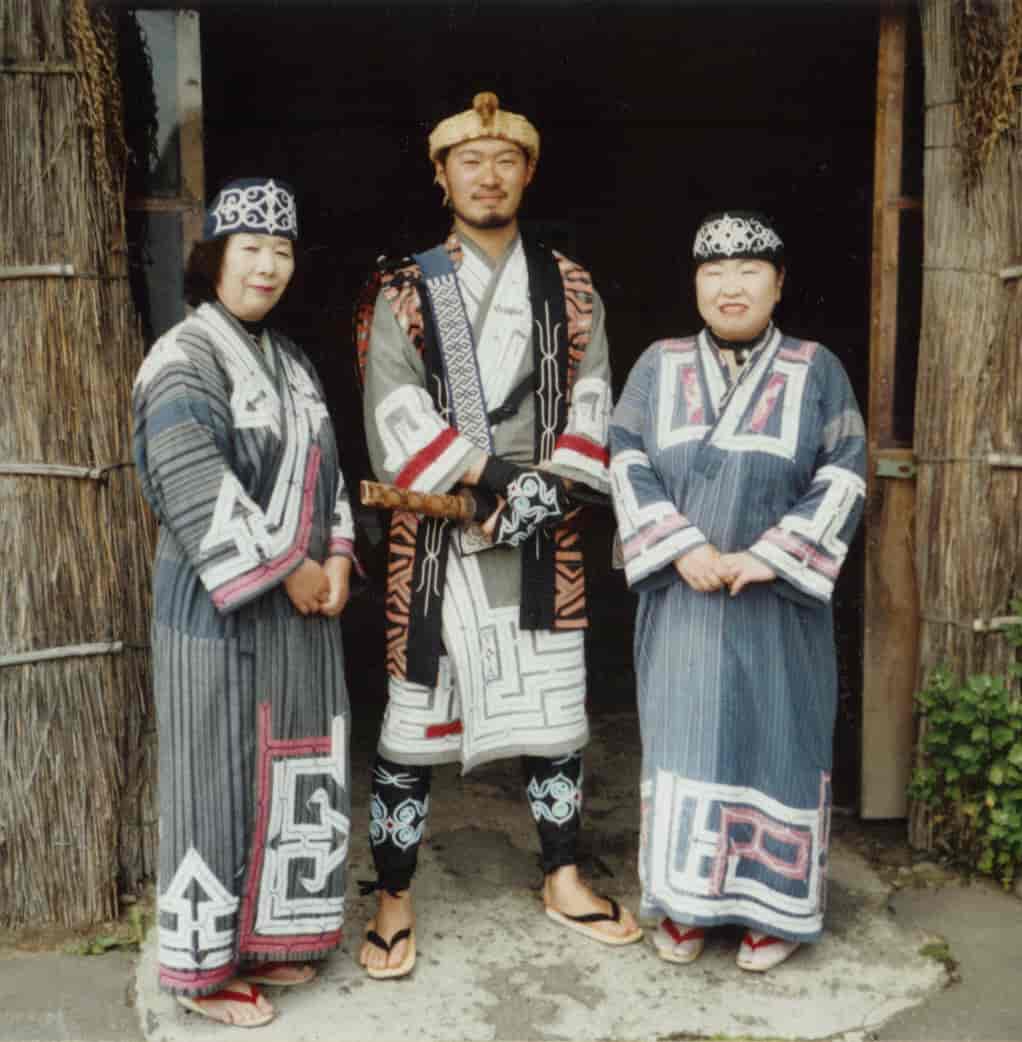 Tre ainoer (en mann og to kvinner) står oppstilt i tradisjonelle klær - lange kapper med broderier.