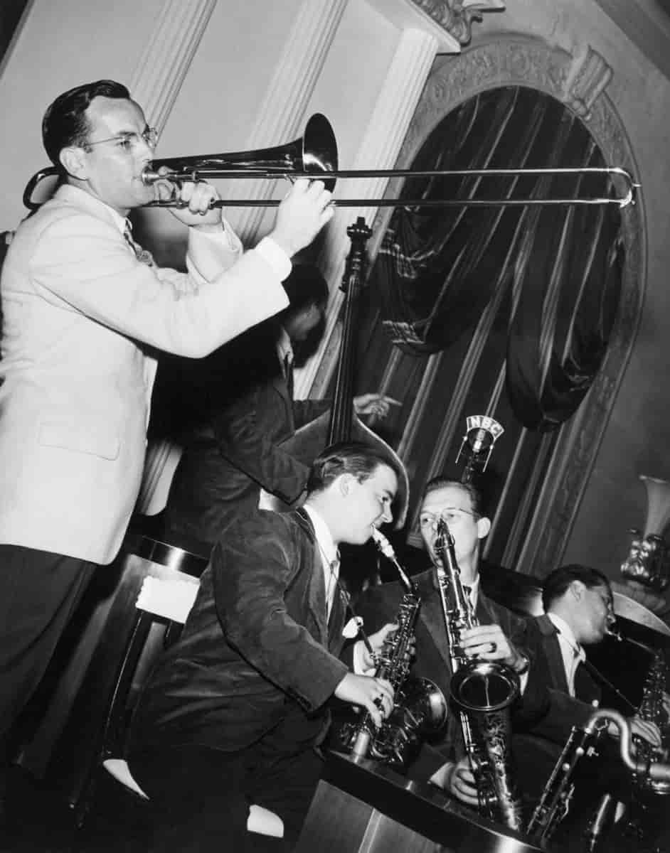 Svart/hvitt bilde av Glenn Miller som spiller på tromone. Til høyre for ham ser man flere musikere, blant annet en som spiller kontrabass og tre som spiller saxofon.