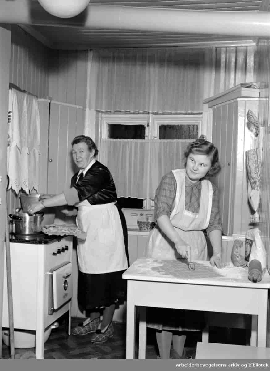 Svart-hvitt foto av to kvinner, en ung og en eldre, på et lite kjøkken. Begge har på forkler. Den unge kvinnen skjærer ut deig i et mønster. Den eldre står ved en gammeldags komfyr og skal til å løfte opp en kjele. På komfyren står det også et brett med bakst. 