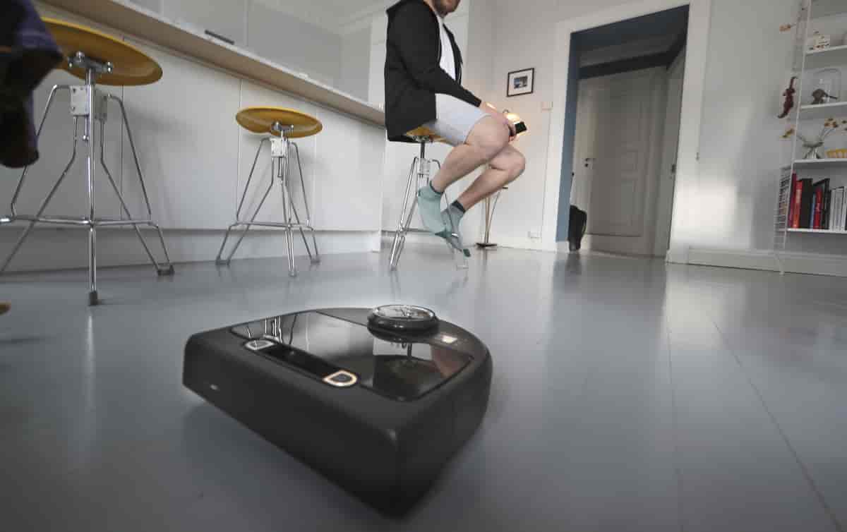 En svart, rund robotstøvsuger på et gulv. I bakgrunnen sitter det en person og ser inn i en mobiltelefon.