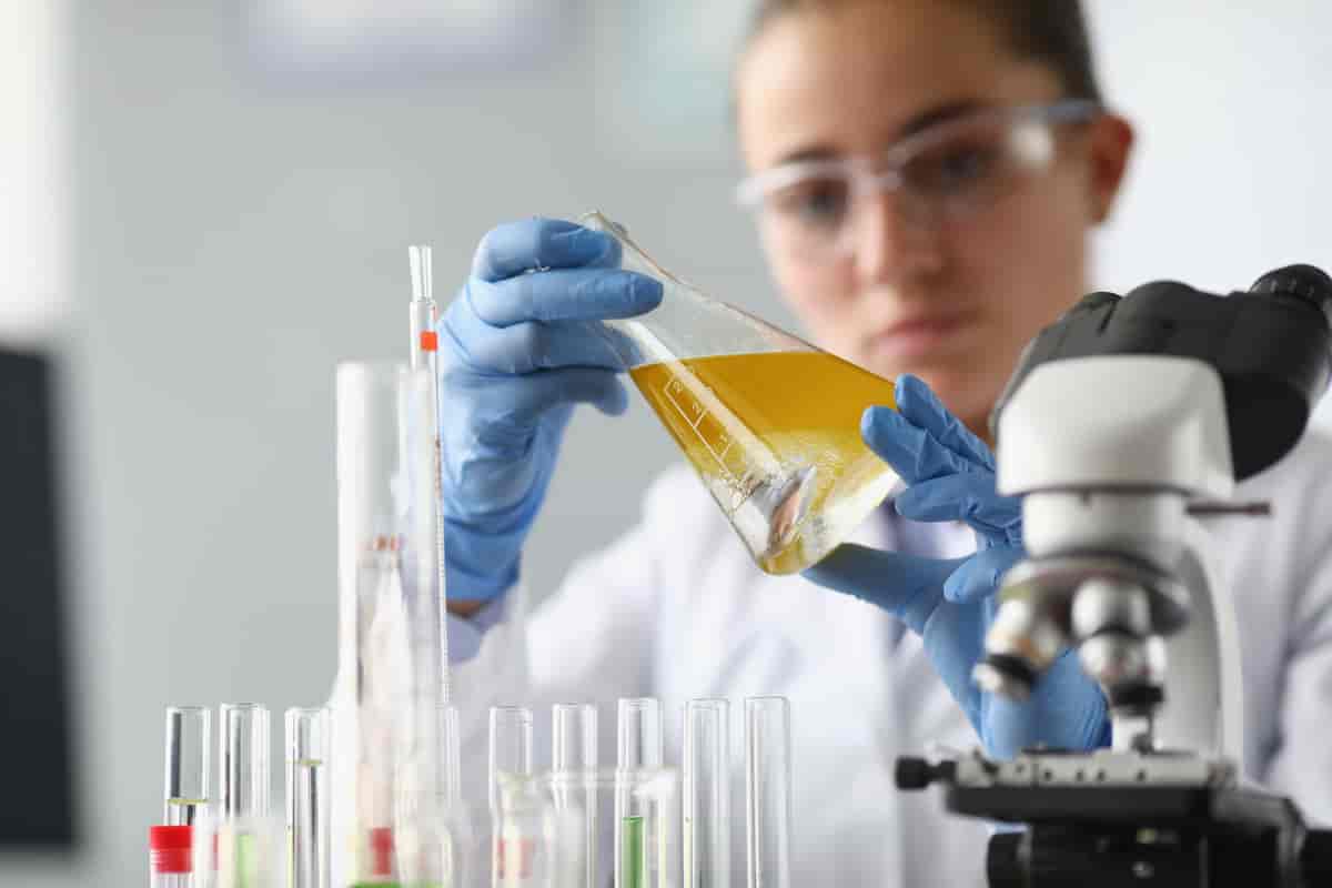 En kvinne med vernebriller som holder et reagensglass i et laboratorium. Det er flere reagensrør med ulik høyde foran henne. Til høyre i bildet er det et mikroskop.