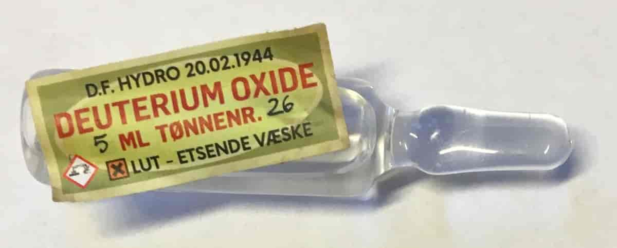 En ampull med klar væske i. Utenpå er det en merkelapp hvor det står "Deuterium oxide. Lut - etsende væske".
