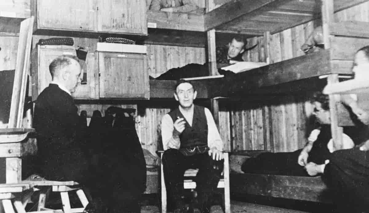 Sort-hvitt fotografi av et lite rom med køyesenger. Professoren sitter på en stol i midten. En annen mann sitter på en benk til venstre. I køyesengene ligger flere andre fanger og hører på.