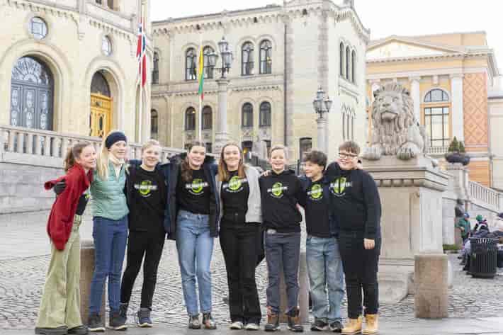 Foto av åtte barn ved den ene løvestatuen utenfor Stortinget. Seks av dem har gensere med logoen til Miljøagentene på brystet.