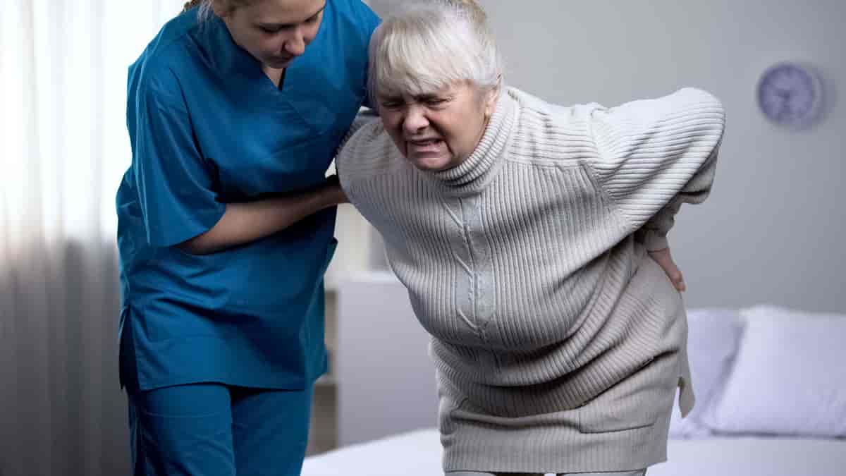 En eldre dame reiser seg opp og tar seg til hoftene. Hun støttes av en sykepleier. 