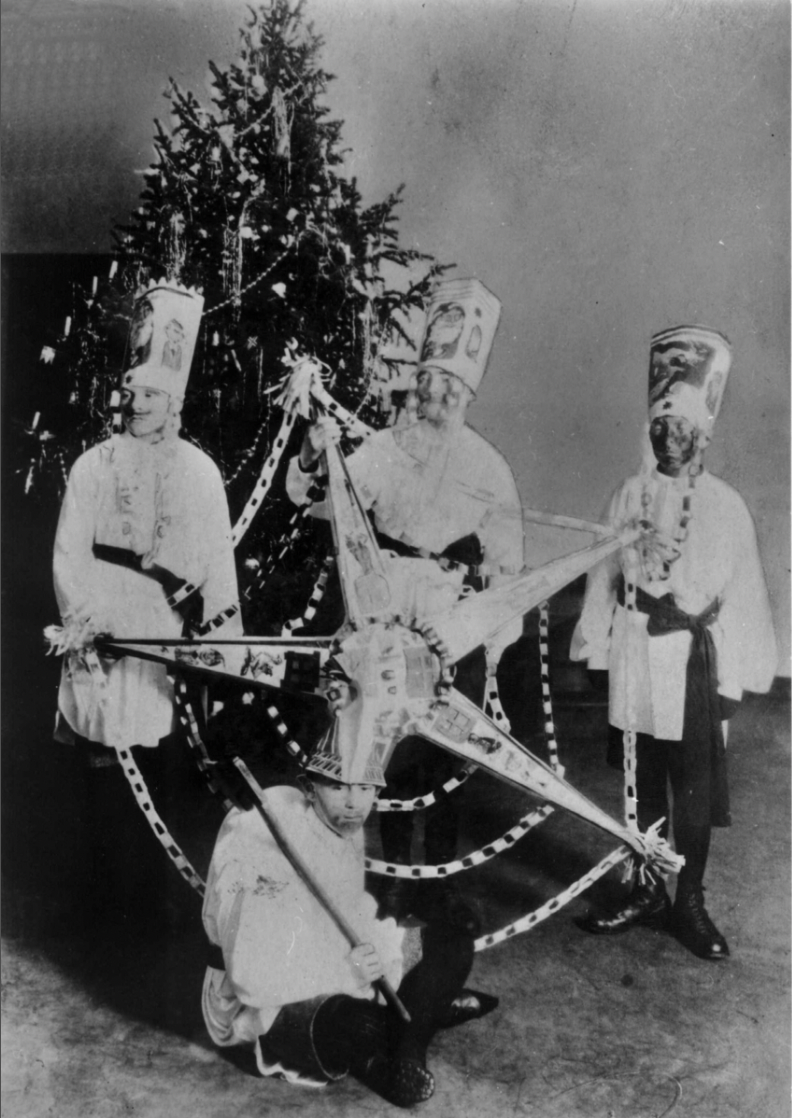 Svart-hvitt foto av fire gutter foran et juletre. Tre av guttene er kledd ut med som de tre vise menn med hatter av papp og skjegg sminket på. Foran dem sitter en gutt med en hatt laget som en stor stjerne. 