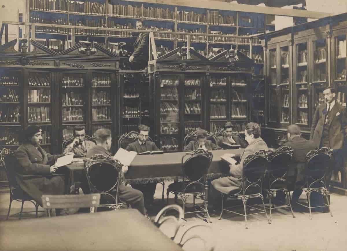 Sort-hvitt fotografi fra biblioteket. En gruppe menn sitter rundt et langbord og leser. I bakgrunnen har en bibliotekar klatret opp i en høy bokhylle.