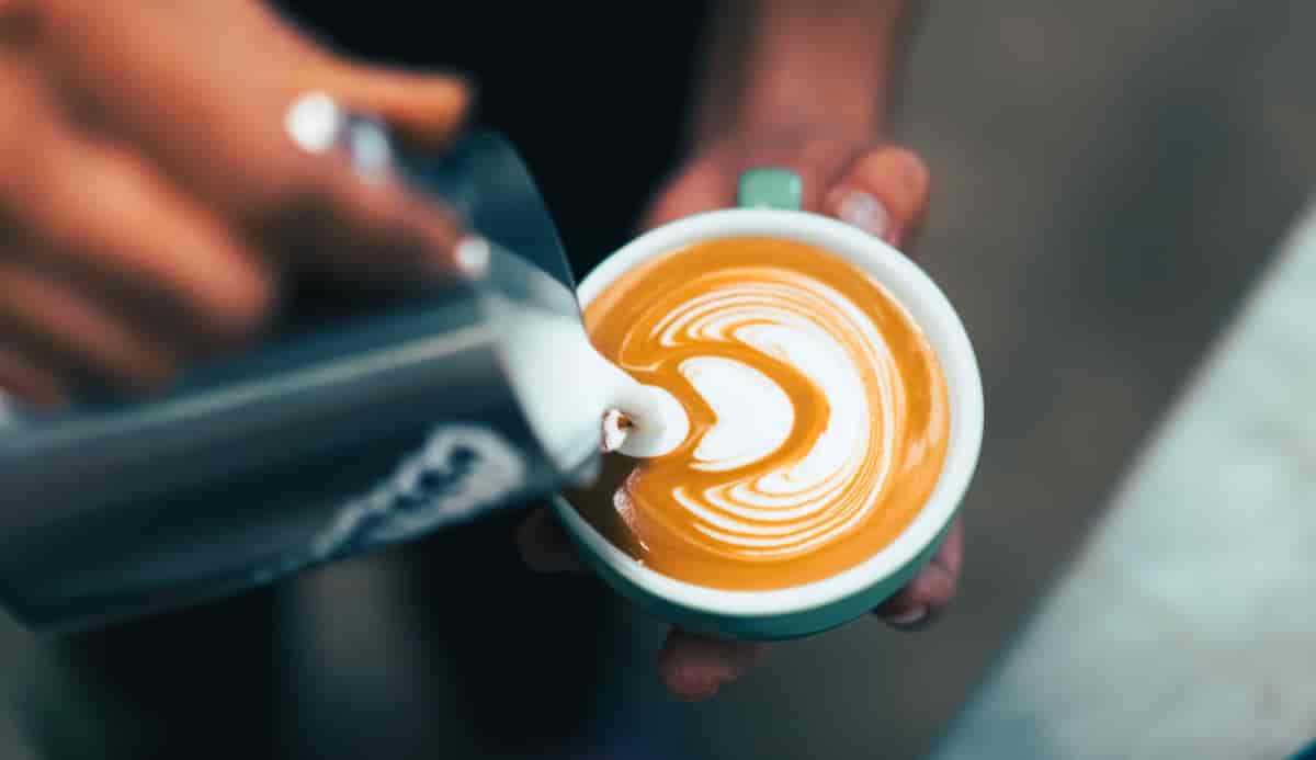 Nærbilde av hender som heller melkeskum fra en liten stålmugge oppi en kopp med kaffe. 