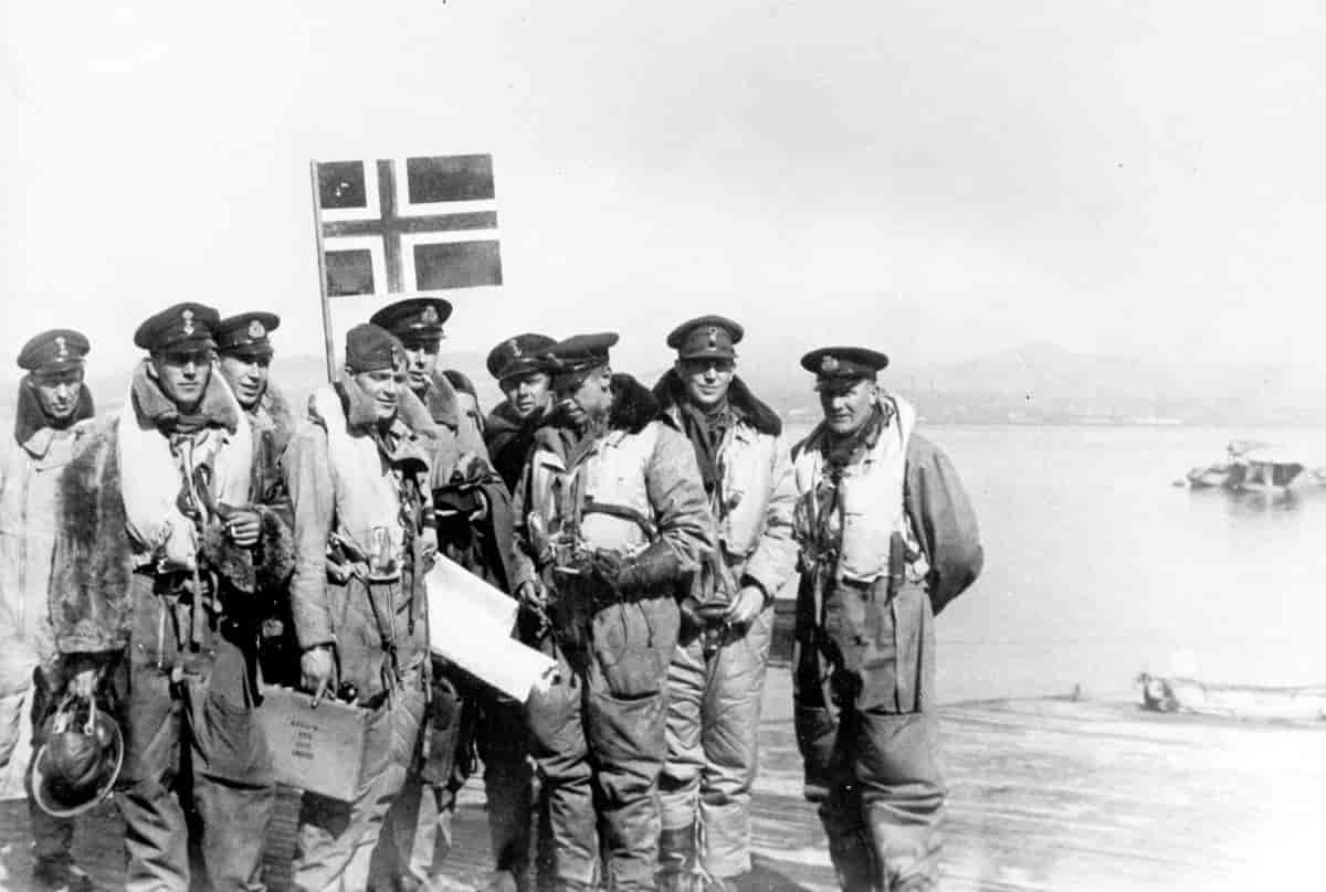 En flokk soldater står ved vannet. I bakgrunnen ser man det norske flagget.