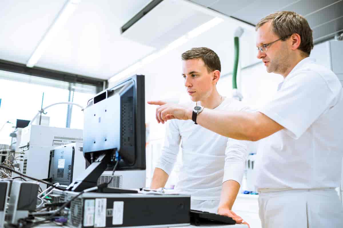 To menn i hvite klær som står foran en datamaskin og ser konsentrert inn i skjermen.