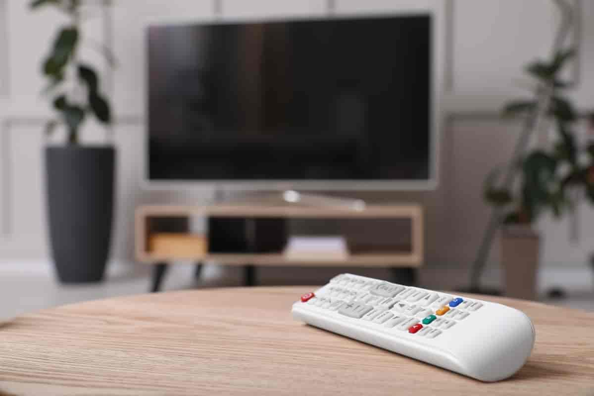 En hvit fjernkontroll med mange knapper som ligger på et stuebord. I bakgrunnen er det en TV.