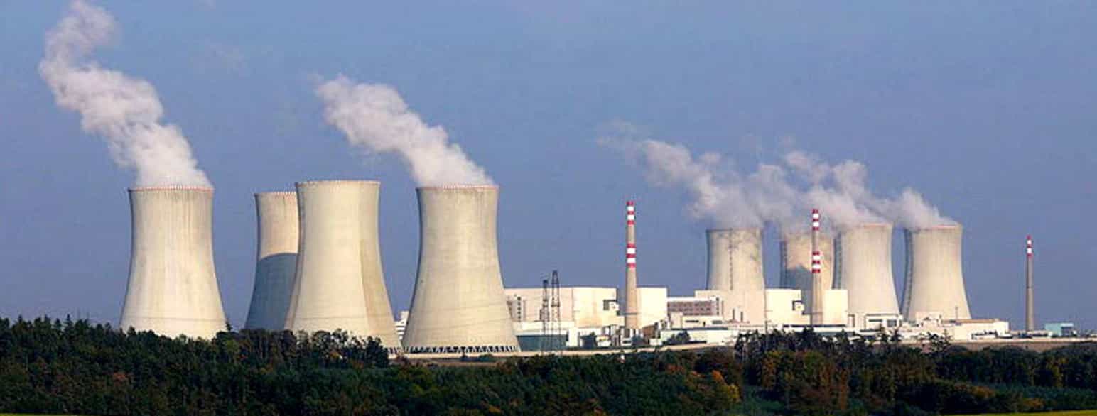 Et kjernekraftverk med store piper i grå betong. Det ryker ut av pipene. I midten av bildet, mellom pipene, ser man ulike fabrikksbygninger.