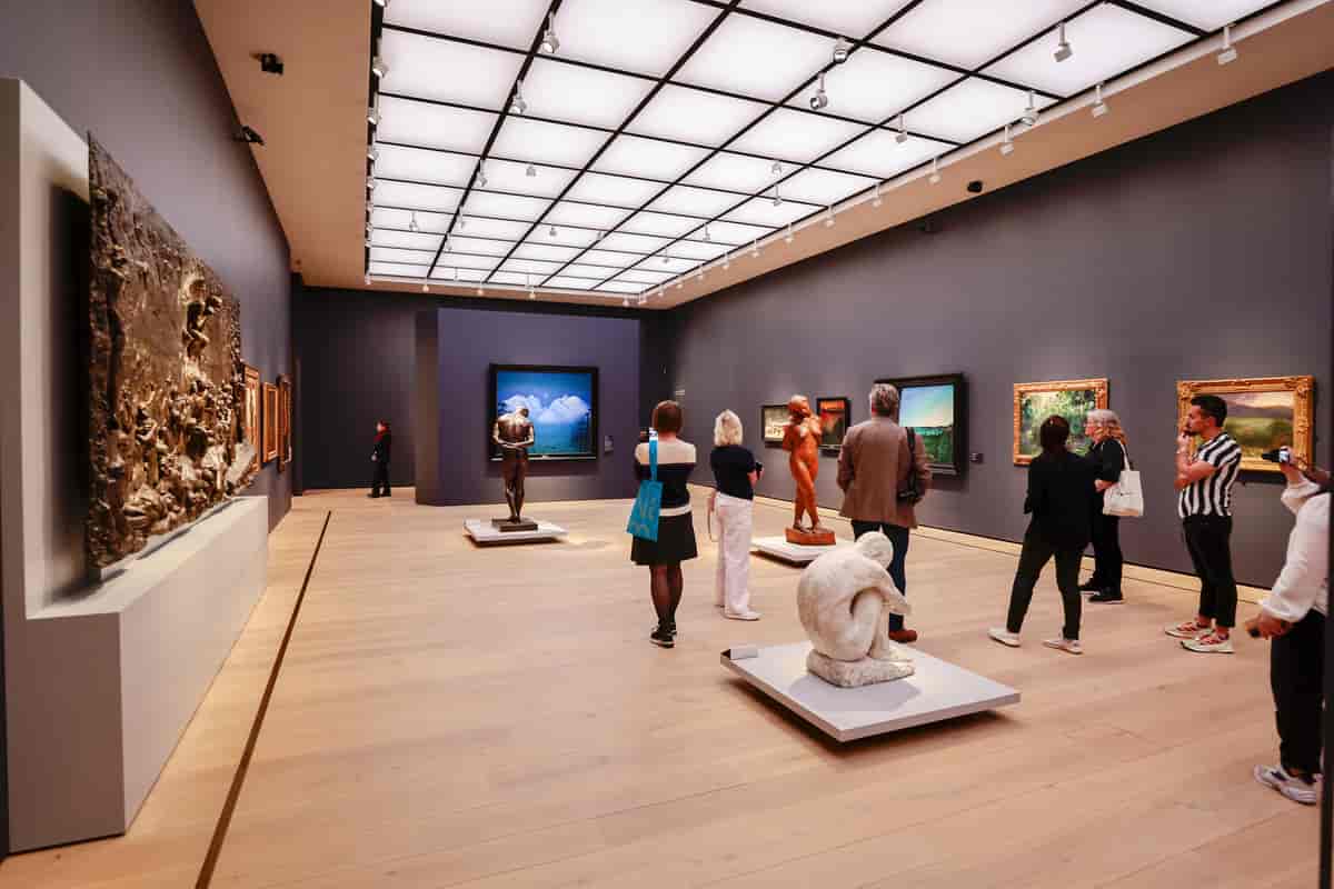 Et rom med malerier og et relieff på veggene. På gulvet står tre skulpturer som forestiller mennesker. I rommet er det også publikum.
