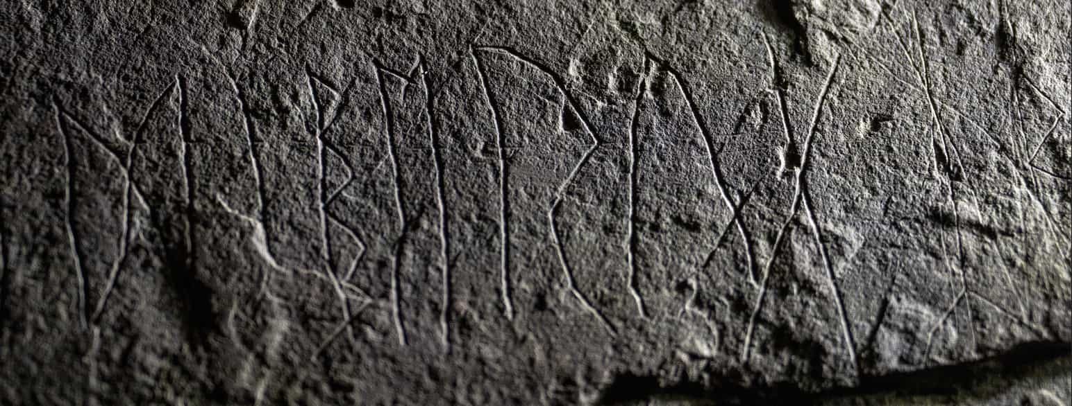 Nærbilde av en grå runestein med runer risset inn.