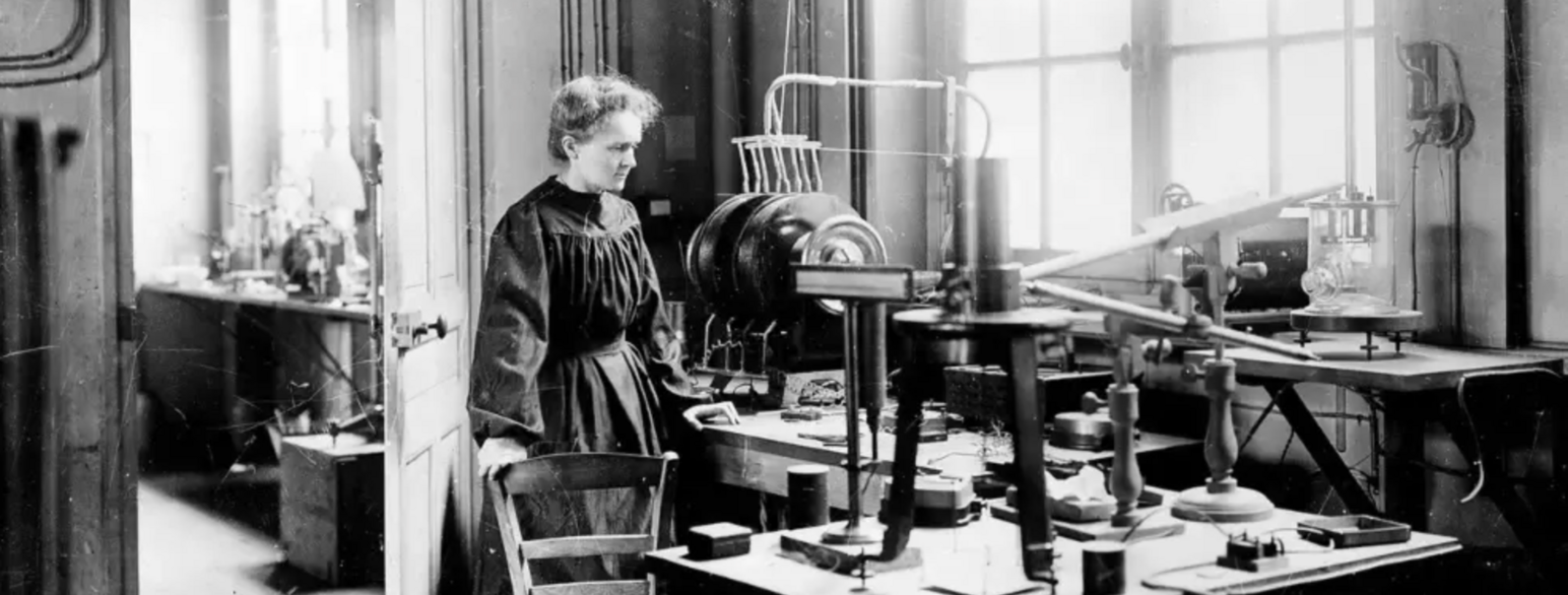 Svart/hvit foto av Marie Curie ved et arbeidsbord i et laboratorium