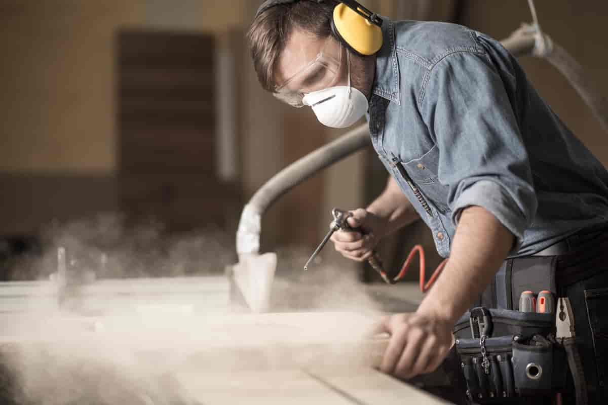 En mann med hørselvern, støvmaske og briller arbeider med bråkete verktøy