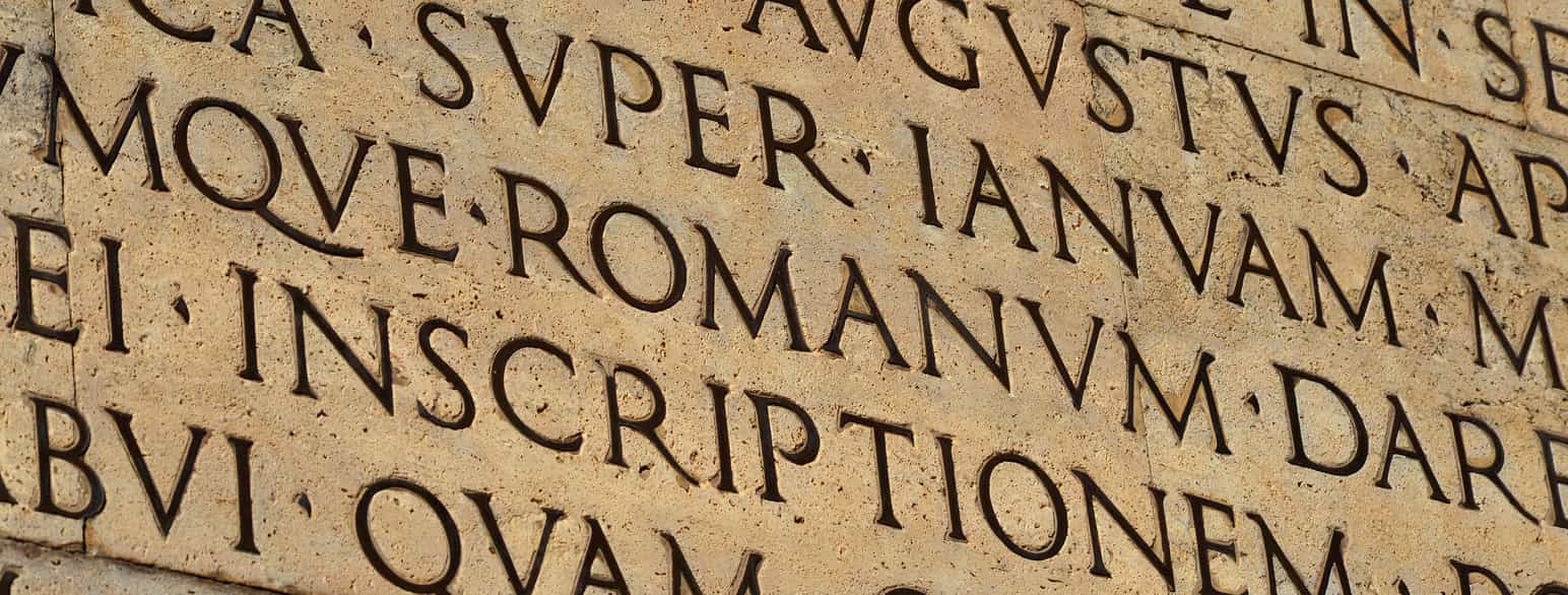 Rissede bokstaver på latin på en gul murvegg.