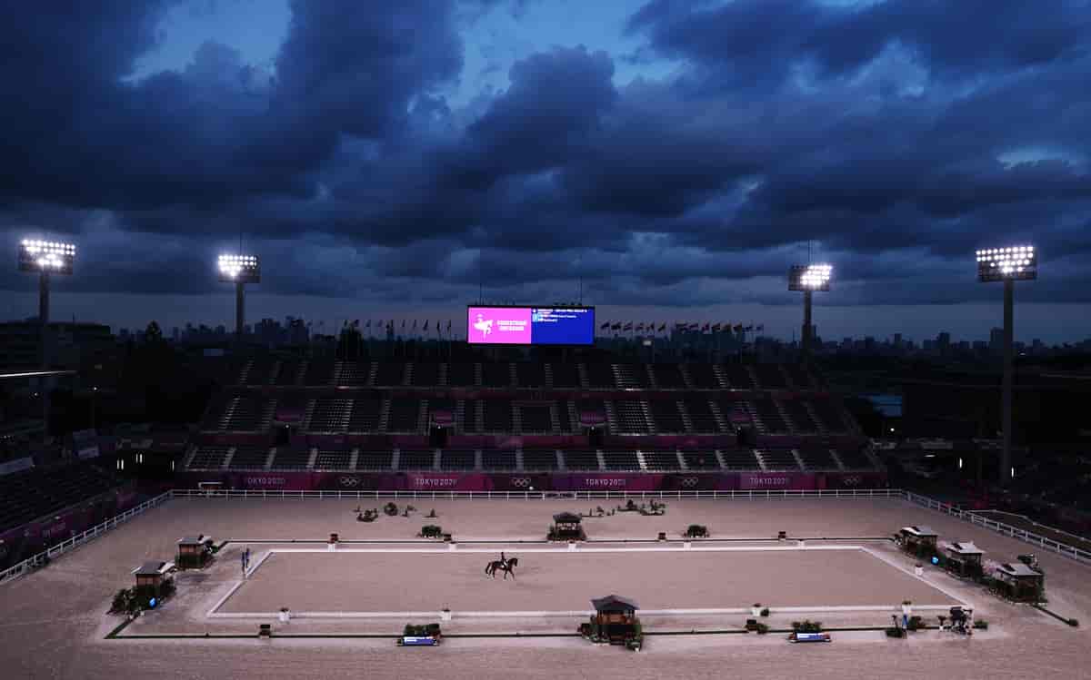 Foto av en arena, der underlaget er sand, og en rytter rir på en hest innenfor et avgrenset område. På den ene langside er det en tribune. 
