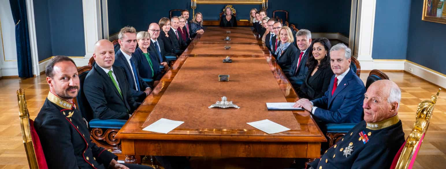 Foto fra statsråd med kronprins Haakon og kong Harald nærmest kamera. Nedover på hver side av et langt langbord sitter regjeringsmedlemmene. 