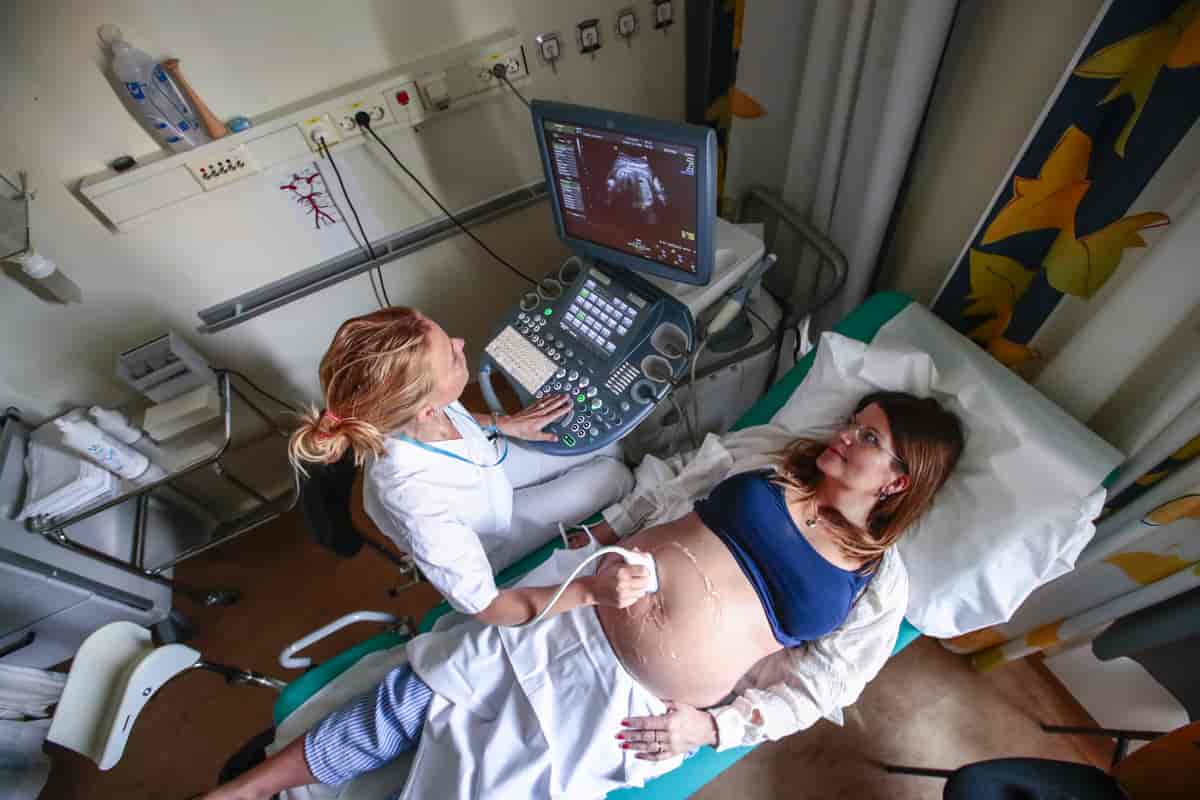 Legen o den gravide er på et sykehusrom. Legen er kledd i en hvit uniform og bruker en maskin for å se inn i magen på den gravide. Bildet av babyen ses på en skjerm.