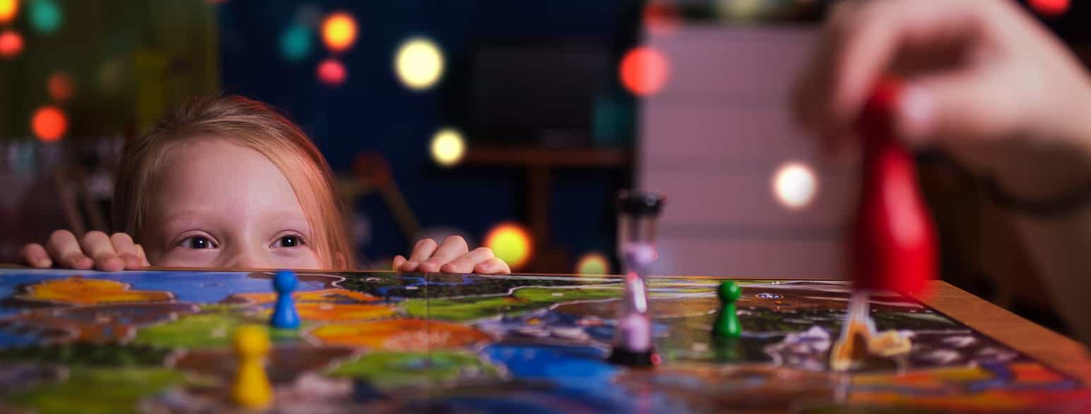 Foto av spillbrett med et timeglass og brikker i forskjellige farger. Et barn holder hendene på spillbrettet og stikker panne og øyne opp over bordkanten.