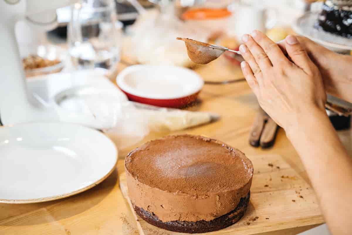 Foto av to armer som drysser brunt pulver over en brun kake. I bakgrunnen er det forskjellige fat og boller og kjøkkenutstyr.