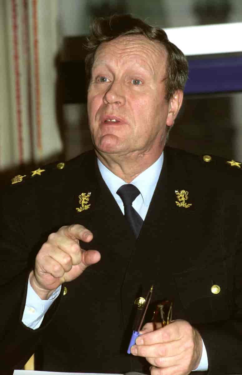 Mann i svart politiuniform med gulldetaljer og slips. Holder et par briller i den ene hånden, og peker foran seg med den andre. 
