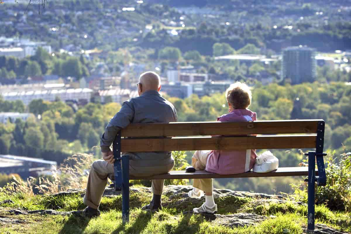 To eldre mennesker, en kvinne og en mann, sitter på en benk og ser utover noe landskap og litt av en by.