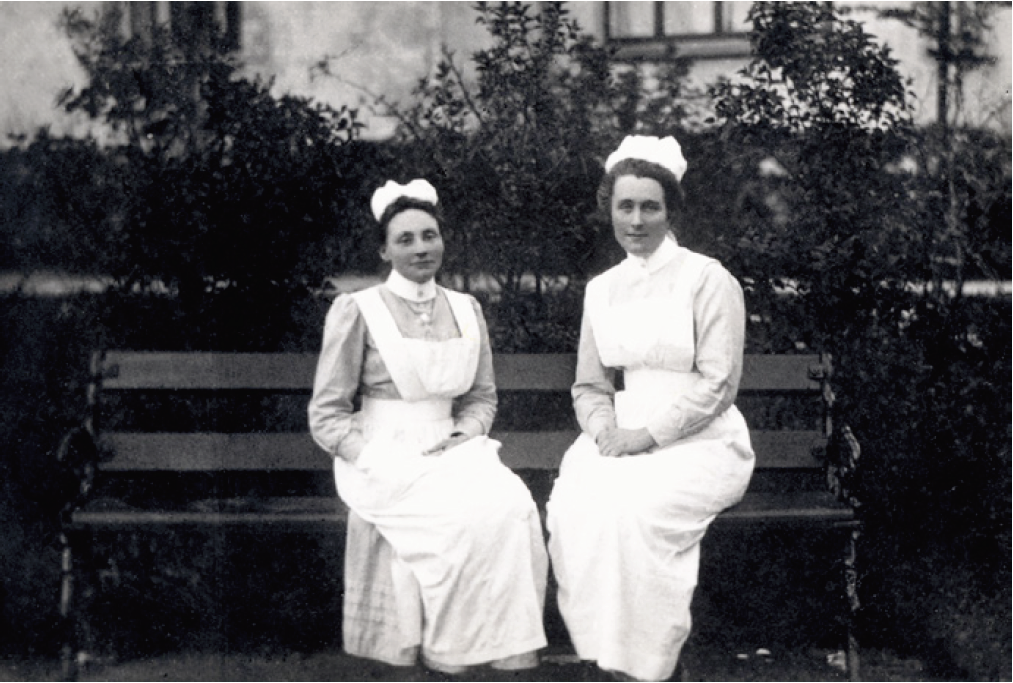 De to kvinnene sitter i sykepleiertøy på en benk utendørs.