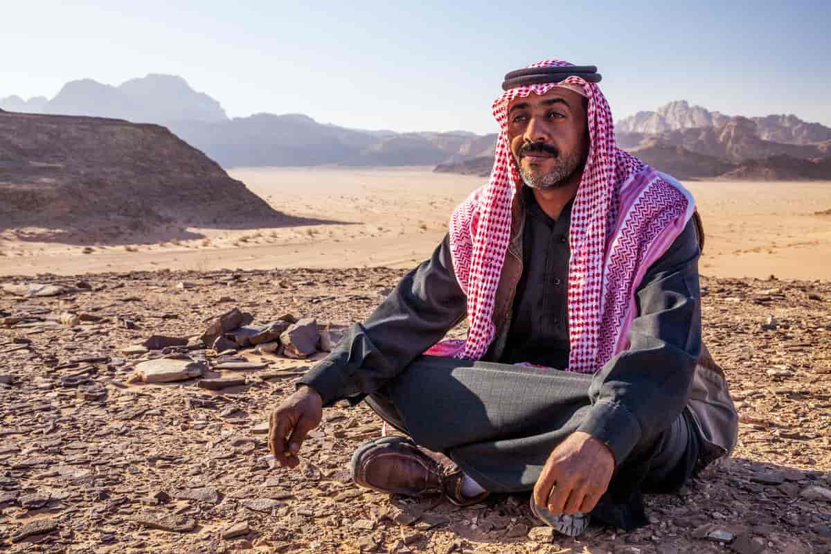 Mann sitter med bena i kryss på bakken, ørken og fjell-landskap i bakgrunnen. Han har et rødt og svart palestinaskjerf på hodet og over skuldrene, med to svarte ringer på toppen av hodet som holder det på plass. 