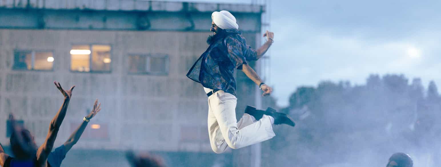 Fargefoto av danseren Partap Singh. Bildet er tatt under en konsert, mens danseren er midt i et hopp. Knærne er bøyd. Han er  praktiserende sikh og har på turban som sikher bruker. Til venstre i bildet ser man armene fra noen i publikum som strekker seg i været. 
