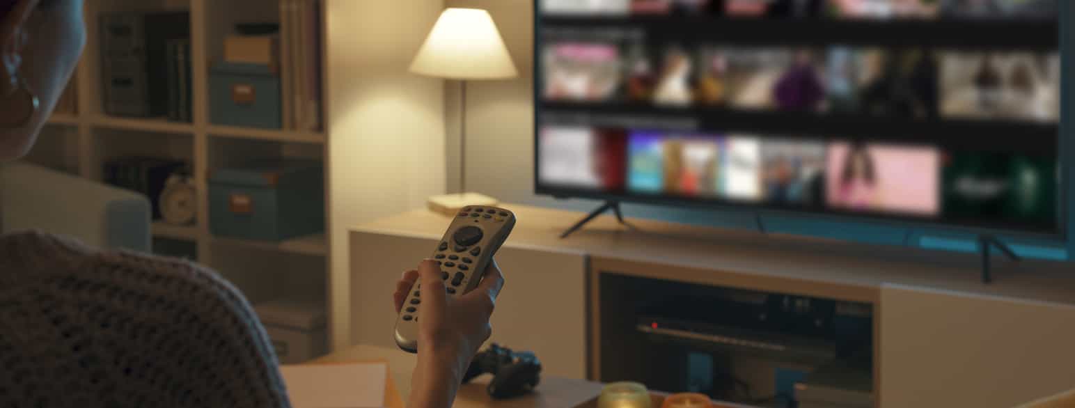 En kvinne bakfra som holder en fjernkontroll i hånden. I bakgrunnen skimter man en TV som står på et TV-bord.