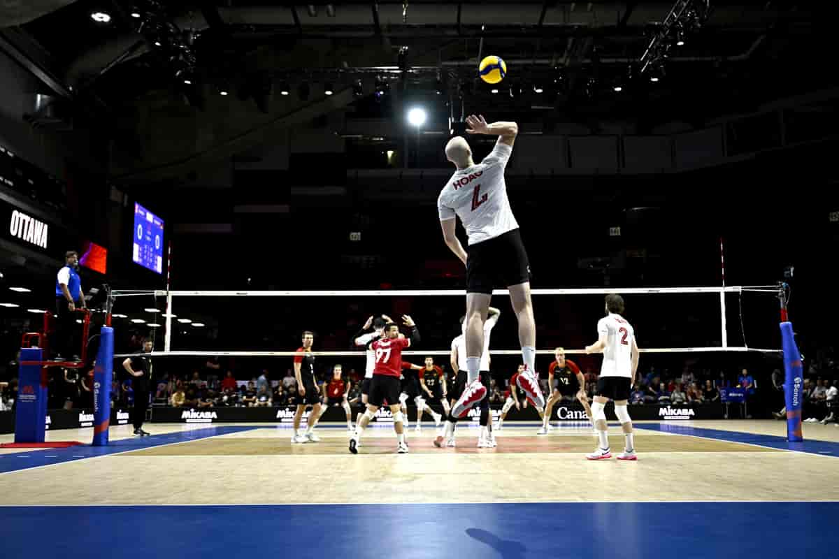 Foto av kamp på volleyballbane. Spilleren som server er i et hopp ganske høyt over gulvet. Den høyre hånden hans er på vei til å slå ballen som er i lufta over ham igjen.