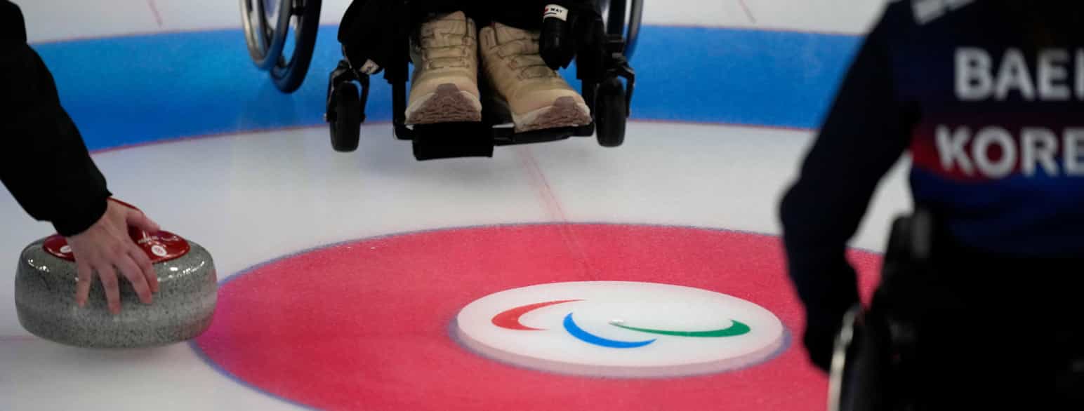 Foto nærbilde av «boet» på en curlingbane med logoen til paralympics i midten. Anskåret er føttene og det nederste av fotbrettet til én spiller, og ryggen, armen og hjulet på rullestolen til en annen.