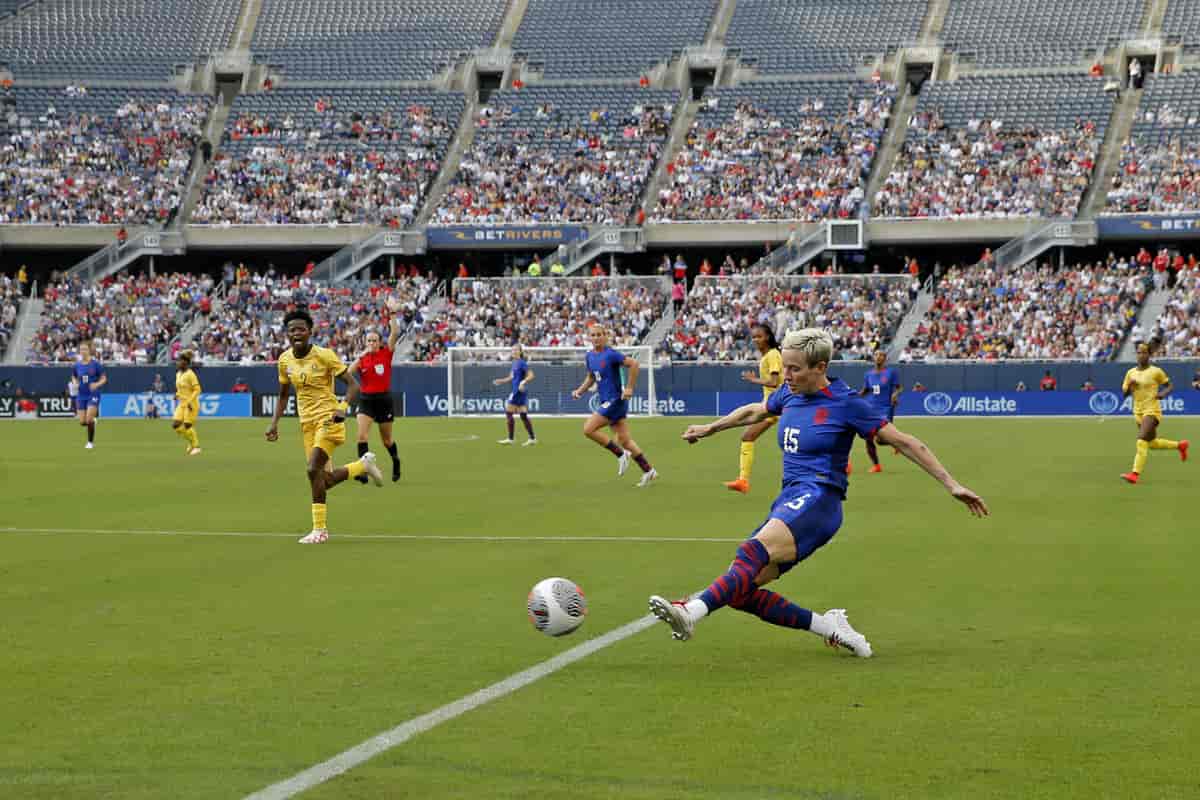 Foto fra fotballkamp. Rapinoe er nærmest kamera på vei mot å sparke ballen med venstre fot. Spilleren som er bak og nærmet henne er Sør-Afrikas nummer 2. 