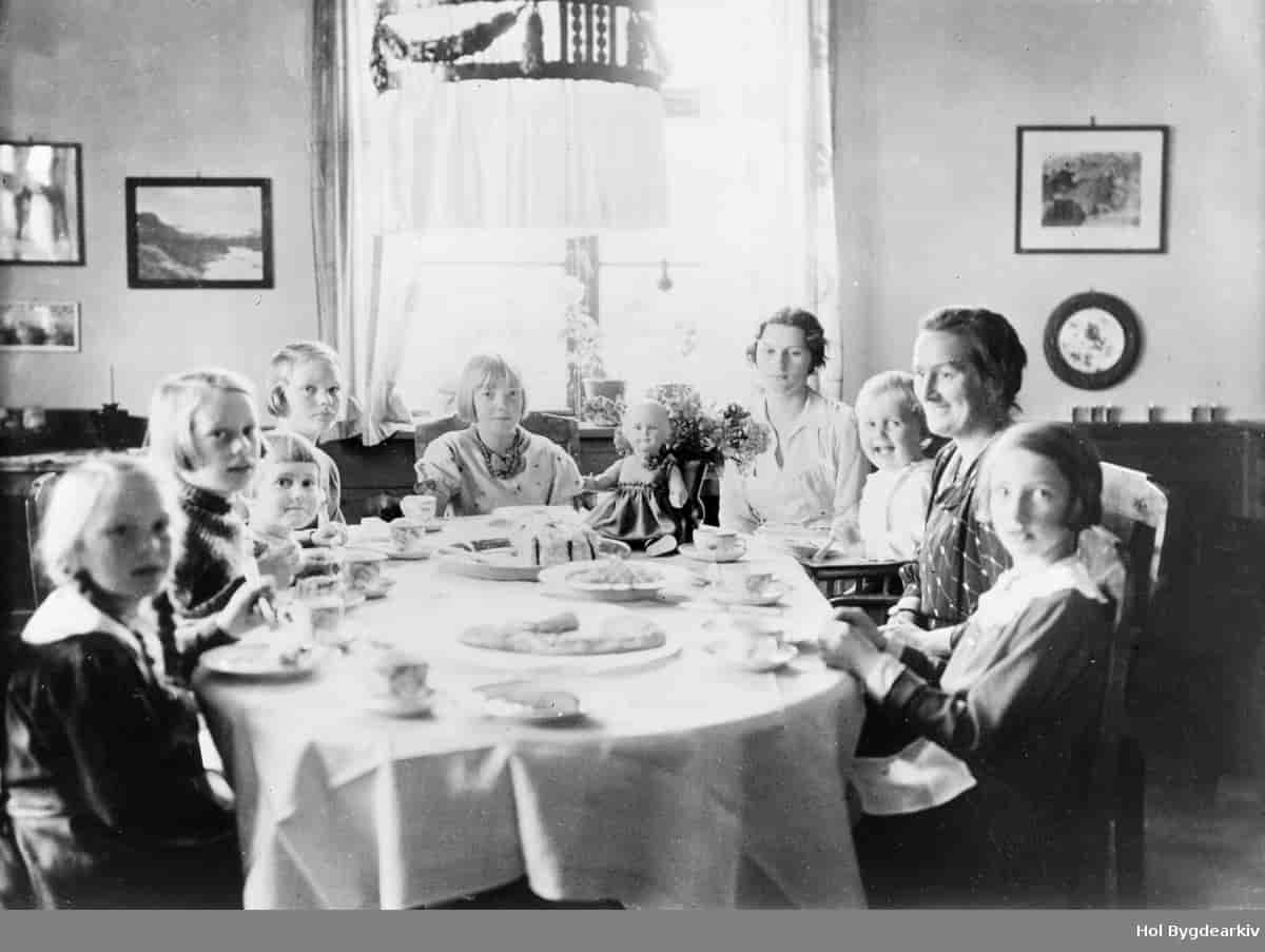 Et svart-hvitt foto av åtte barn og to voksne rundt et bord. Det er hvit duk på bordet, og dekket på med små tallerkener og kaffekopper. Det står to kaker på bordet.