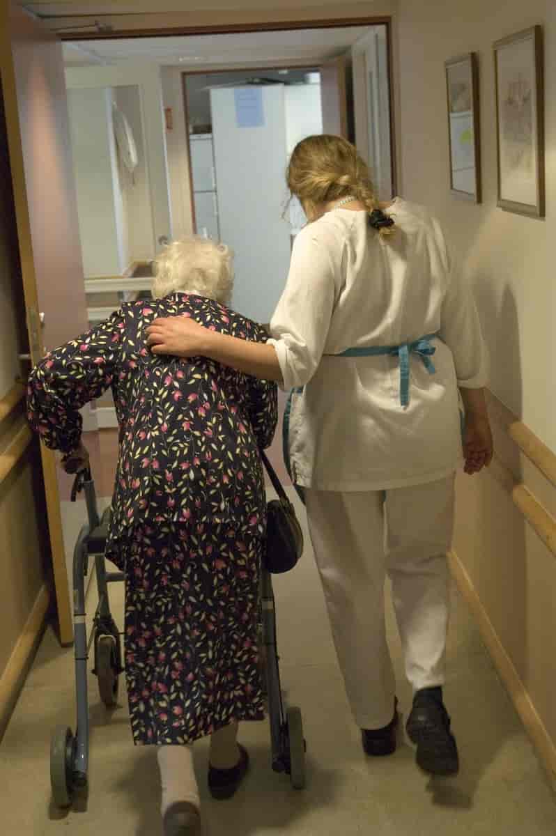 En eldre kvinne bakfra som støtter seg til en gåstol. Ved siden av henne går en sykepleier i hvit uniform.