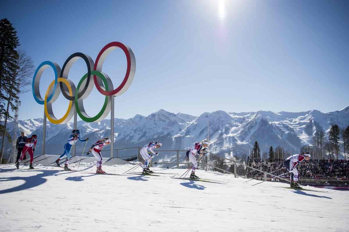 Foto fra langrennsløype i strålende sol. Sju løpere går etter hverandre. Bak løperne og OL-ringene er det en rekke høye, snødekte fjell.