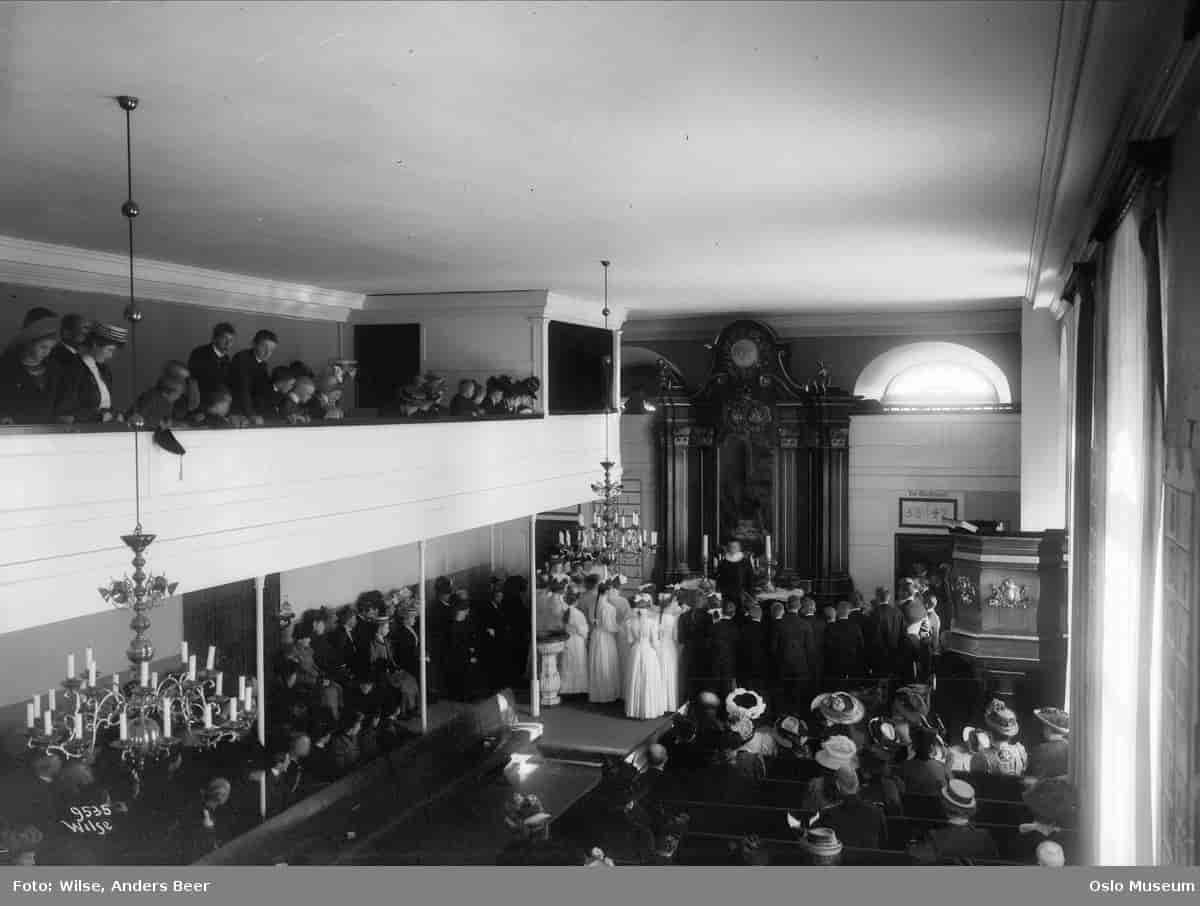 Sort-hvitt fotografi av kirkerommet med konfirmanter stående framme ved alteret hos presten.