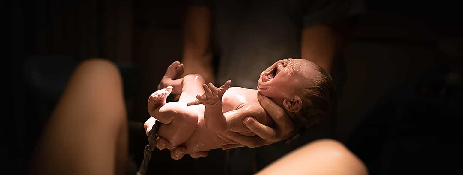 Foto av nyfødt baby. Babyen er naken og gråter. Babyen er litt klissete og våt, og man kan se navlestrengen fra magen. En voksen holder babyen, og man kan se beina til mora. 