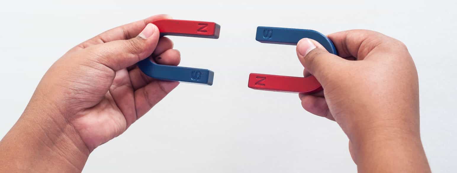 To hender som holder magneter formet som hestesko i hver hånd. Halvparten av hver magnet er blå, der står det S. Halvparten av hver magnet er rød, der står det N. Magnetene er holdt slik at den blå siden på den ene magneten står mot den røde siden på den andre.