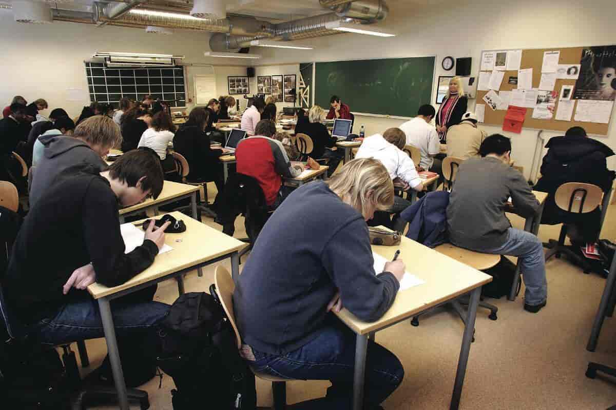 Mange elever som sitter og skriver ved pulten sin. Foran i klasseromet sitter det en lærer, og det står en lærer ved siden av tavlen.