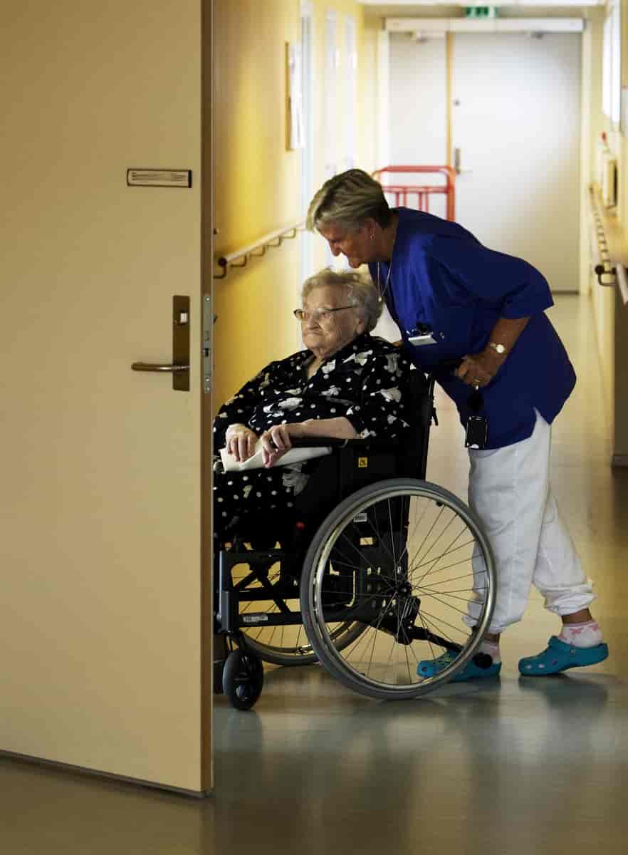 En eldre kvinne i rullestol blir hjulpet inn på rommet av en sykepleier.