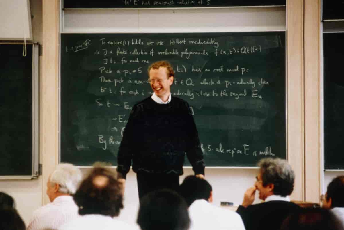 En smilende Andrew Wiles står foran en tavle med mange matematiske symboler på. Foran ham er det flere menn som sitter i salen. 