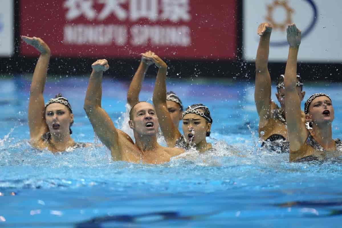 Foto av synkronsvømmere med hodet og skuldrene over vann og den ene armen løftet. Det er fem kvinner og én mann.