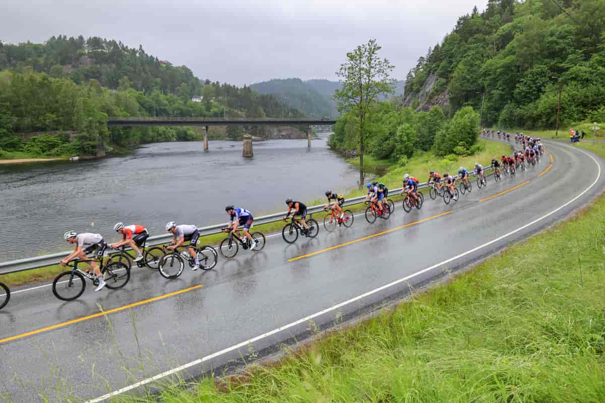 Foto av syklister på en asfaltert vei ved siden av ei elv med en bro over. Bakken er våt etter regnvær.