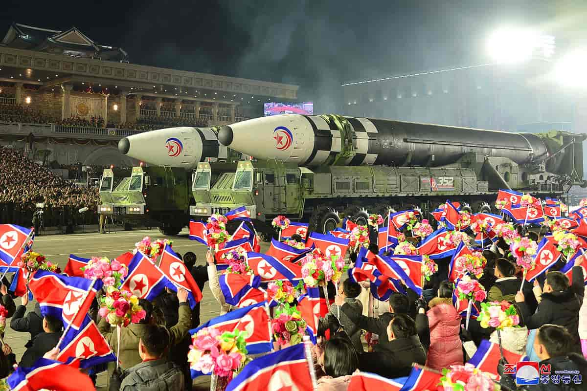 Foto av en står åpen plass. En folkemengde med nord-koreanske flagg og to militære lastebilder med to kjempestore raketter. I bakgrunnen en stor bygning med en balkong hvor det er masse folk i militæruniformer.