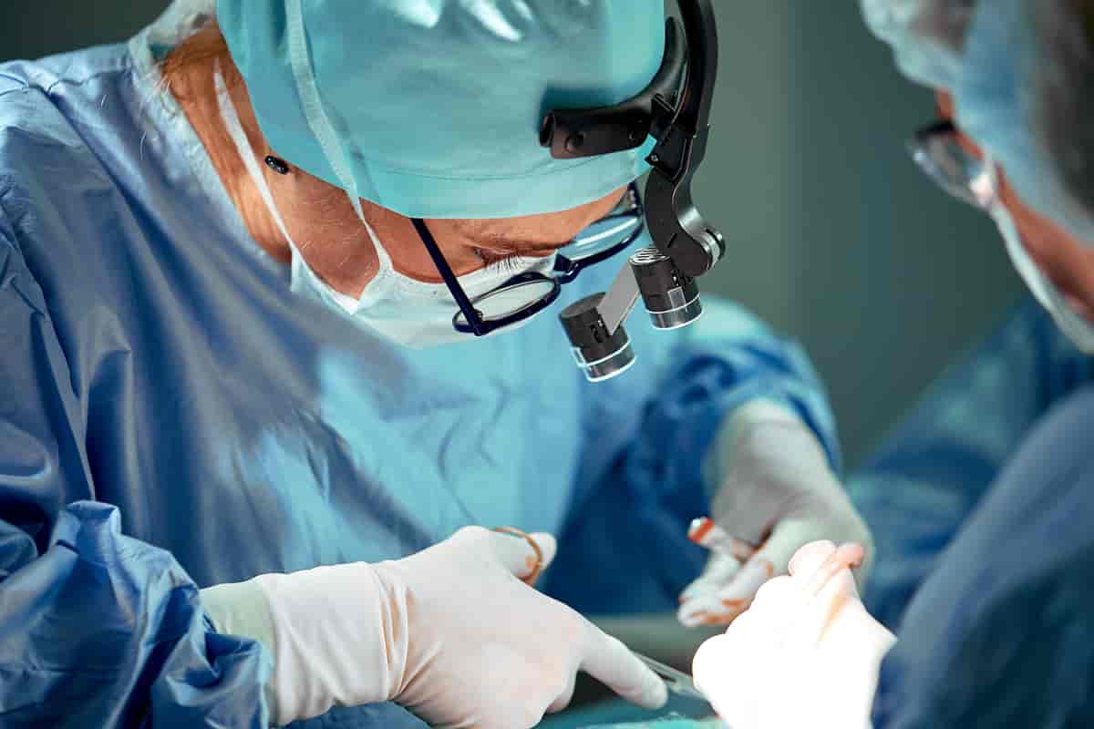 Kirurg i sjukehusklede, hårnett og munnbind arbeider med ei saks på ein pasient. Hen har på seg hanskar med litt blod på, og to par briller så hen kan sjå godt.