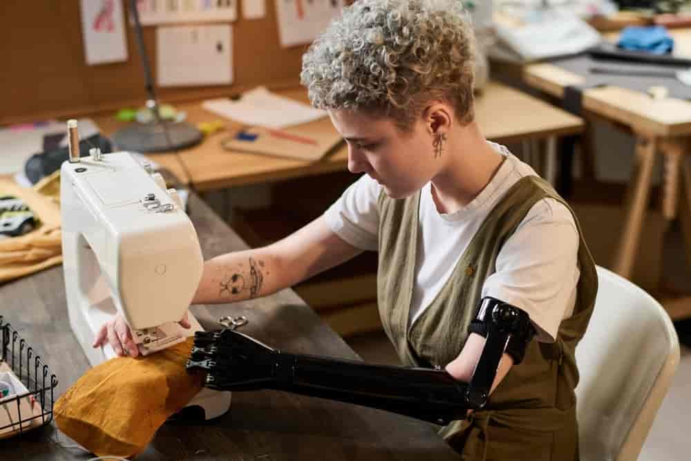 Ung kvinne med armprotese som syr på symaskin. Protesen begynner over albuen og har albueledd og noe som ser ut som en hånd. 