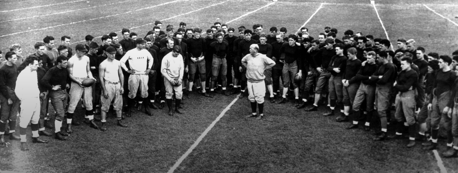 En rekke spillere i amerikansk fotball står samlet rundt Knute Rockne som gir instrukser.